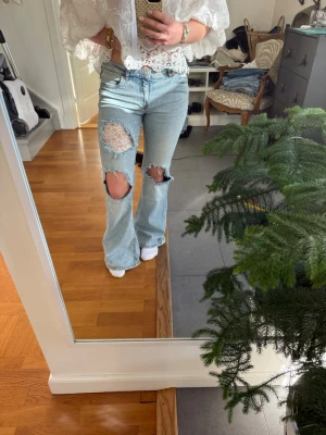 Ljusblå slitna bootcut jeans - Säljer ett par ljusblå bootcut jeans med stora slitningar och hål på båda knäna. Jeansen har hög midja, klassiska fem fickor och är tillverkade i denim. Perfekta för en avslappnad och trendig look.