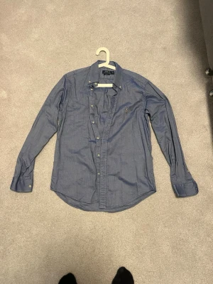 Blå skjorta från Polo Ralph Lauren - Klassisk blå skjorta från Polo Ralph Lauren med broderad logga på bröstet. Skjortan har lång ärm, knappar framtill och en tidlös passform. Perfekt för dig som gillar stilrena och ikoniska plagg.