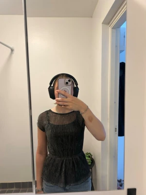 Glittrig svart topp med kort ärm - Snygg svart topp med glittrigt tyg och korta ärmar. Modellen har en markerad midja och lätt utsvängd nederdel. Perfekt för dig som vill sticka ut lite extra. Tunt, lätt material som känns bekvämt mot huden.