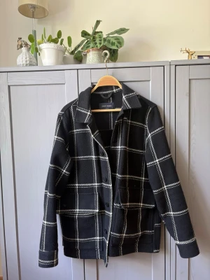 Svart overshirt/jacka med rutigt mönster - Stilren svart overshirt från A days march med vitt rutigt mönster. Klassisk krage, knäppning framtill och två stora fickor. Overshirten har en rak passform och är tillverkad i ett tjockare ull tyg som ger en cool look.