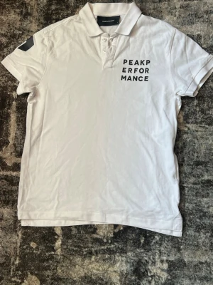 Vit Peak Performance pikétröja - Vit pikétröja från Peak Performance med svart tryck på bröstet. Klassisk krage med knappar och korta ärmar. Perfekt för en clean och sportig stil. Tillverkad i mjuk bomull för maximal komfort.