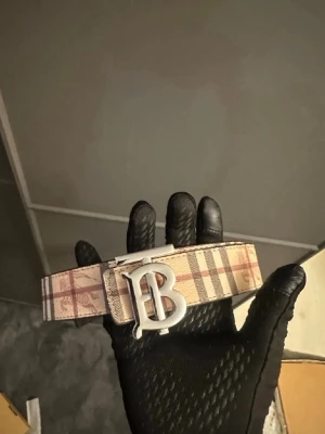 Burberry Bälte - Snyggt bälte från Burberry i beige med klassiskt rutigt mönster och ett stort silverfärgat B-spänne. Perfekt accessoar för att lyfta din outfit med en ikonisk touch. Bältet har en stilren design och passar till många olika looks.
