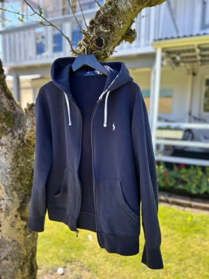 Mörkblå hoodie från Polo Ralph Lauren - Mörkblå hoodie med dragkedja från Polo Ralph Lauren. Klassisk design med vit logga på bröstet, två fickor fram och vit dragsko i huvan. Perfekt för en chill och stilren look. Mjuk bomullskänsla och ribbade muddar.