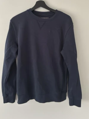 Mörkblå sweatshirt med rund hals - En stilren mörkblå sweatshirt med rund halsringning och långa ärmar. Tröjan har ribbade muddar vid ärmslut och nederkant, perfekt för en avslappnad och enkel look. Passar till jeans eller joggers.