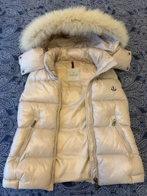 Moncler Beige dunväst med pälskrage   - Snygg beige dunväst från Moncler med avtagbar huva och fluffig pälskant. Västen har två dragkedjefickor framtill, quiltad design och klassisk Moncler-logga på bröstet. Perfekt för lager-på-lager, 1 av 1 kvalitet.