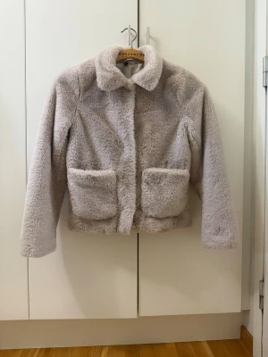 Ljusgrå pälsjacka från H&M - Supermjuk och fluffig ljusgrå pälsjacka från H&M. Jackan har klassisk krage, två stora fickor framtill och stängs med dolda knappar. Perfekt för dig som vill ha en stilren och cozy look under kyliga dagar.