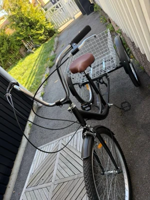 Svart trehjulig cykel med korg - Säljer en svart trehjulig cykel med stor metallkorg bak. Cykeln har brunt sadel, högt styre och robusta däck. Perfekt för att transportera saker eller handla. Metallram och korg i silverfärgat stål.