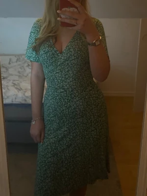 Grön blommig omlottklänning - Snygg grön omlottklänning med små vita blommor och v-ringad hals. Klänningen har korta ärmar och en feminin passform som framhäver midjan. Perfekt för dig som gillar en fräsch och somrig stil.