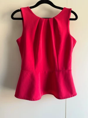 Rosa peplumtop med dragkedja - Snygg röd peplumtop med rund halsringning och markerad midja. Toppen är ärmlös och har en synlig dragkedja i ryggen som ger en cool detalj. Perfekt att styla med jeans eller kjol för en trendig look. Endast provad katt finns i hem!