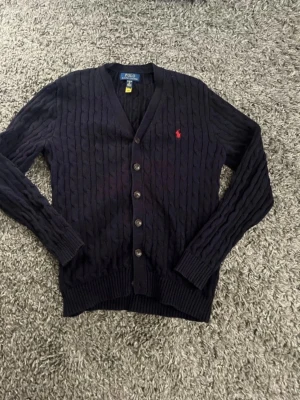 Polo Ralph Lauren Cardigan - Mörkblå kabelstickad kofta från Polo Ralph Lauren med V-ringning och knappar framtill. Klassisk röd logga broderad på bröstet. Långa ärmar och ribbade muddar. Perfekt för lager-på-lager och snygg till jeans.