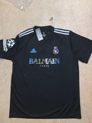 XL. Svart Real Madrid x Balmain tröja - Säljer den för köpte den till min bror på födelsedags present men han supportar antagligen inte real Madrid Snygg svart fotbollströja från Adidas i samarbete med Balmain och Real Madrid. Tröjan har vita detaljer, klubbmärke, holografisk Balmain-logga och nummer 7 med Ronaldo på ryggen. Korta ärmar och sportig passform.