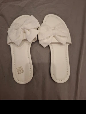 Vita tofflor med stora rosetter - Snygga vita tofflor med bred rem och stora dekorativa rosetter framtill. Tofflorna har en klassisk öppen design och är tillverkade i ett mjukt material som känns bekvämt på foten. Perfekta för att chilla hemma eller på semestern.