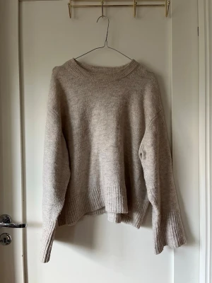 Beige stickad tröja - Mysig beige stickad tröja med oversized passform och breda ribbade muddar vid ärmslut och nederkant. Tröjan har rund halsringning och är tillverkad i ett mjukt material som ger en avslappnad look. Perfekt för lager på lager under kyliga dagar.
