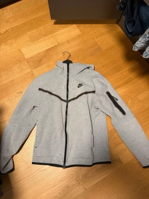 Grå Nike hoodie med dragkedja - Snygg grå hoodie från Nike med hel dragkedja och svart logga på bröstet. Huva och svarta detaljer längs dragkedjan och bröstet. Ficka med dragkedja på ena ärmen. Perfekt för en sportig och avslappnad stil.