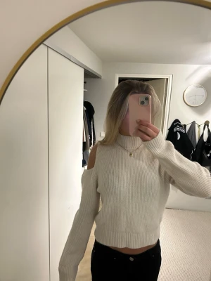 Vit stickad tröja med cut out-detaljer - Trendig vit stickad tröja med cut out-detaljer på axlarna och ribbstickade muddar. Tröjan har en hög hals och en rak, något kortare passform. Perfekt för dig som vill ha en modern och stilren look.