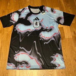 Aston villa special edition  - Cool sport-t-shirt från Adidas i samarbete med KVC Westerlo. Tröjan har ett unikt abstrakt mönster i svart, ljusblått och rött, med klubbens logga på bröstet och Adidas-logga ovanför. Tillverkad i lätt och ventilerande material, perfekt för träning eller match.