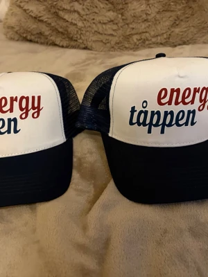 Energytappen truckerkeps blå/vit - Säljer två snygga truckerkepsar från Brandstationen med texten 'energytappen' i rött och blått. Kepsarna är vita framtill med blått nät och böjd skärm. Perfekt accessoar för en cool och avslappnad stil.