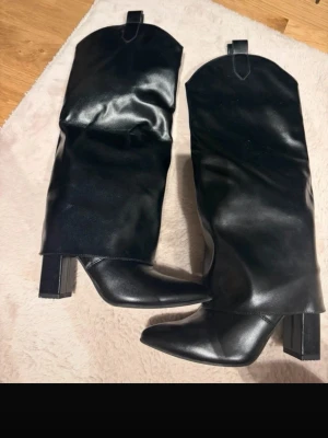 Svarta höga boots med klack - Säljer ett par svarta höga boots med bred klack och spetsig tå. Bootsens skaft är vida och slouchy, tillverkade i glansigt konstläder. Perfekta för att lyfta vilken outfit som helst.