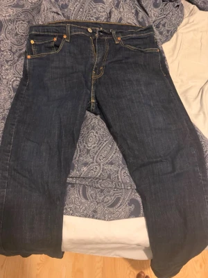 Mörkblå Levi's jeans med raka ben - Säljer ett par klassiska mörkblå jeans från Levi's med raka ben och fem fickor. Jeansen har kontrastsömmar och en ikonisk läderpatch bak i midjan. Tillverkade i slitstark denim, perfekta för en avslappnad och tidlös stil. Storlek 34/32