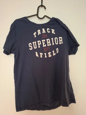 Blå t-shirt från Reaction, tryck - Mörkblå t-shirt från Reaction med vit och röd text på bröstet: 'Track Superior Dept & Field 1994'. Klassisk rund hals och korta ärmar. Skön passform och tillverkad i mjuk bomull, perfekt för en avslappnad stil.
