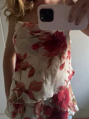 Somrigt blommigt linne med volangkant - Snyggt linne med blommigt mönster i rött och rosa på en ljusbeige bas. Toppen har tunna axelband och volangkant nertill, vilket ger en somrig känsla. Perfekt att matcha med jeans eller kjol.