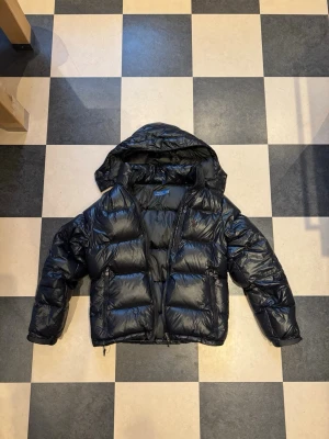 Svart dunjacka med huva från Polo Ralph Lauren - Säljer en svart, glansig dunjacka från Polo Ralph Lauren med stor huva och dragkedja framtill. Jackan har två sidofickor med dragkedja och diskret logga på bröstet. Perfekt för kalla vinterdagar och riktigt snygg streetstil.