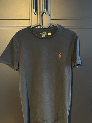 Svart t-shirt från Polo Ralph Lauren - Klassisk svart t-shirt från Polo Ralph Lauren med rund halsringning och korta ärmar. På bröstet finns den ikoniska röda Polo-loggan broderad. Tillverkad i mjuk bomull som känns skön mot huden. Perfekt basplagg för en clean och stilren look. Tröjan är i Slim fit storlek XS men passar även S och är i 7/10 skick 