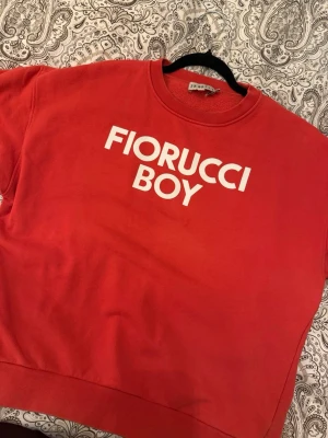 Röd sweatshirt Fiorucci Boy - Säljer en röd sweatshirt från Fiorucci med vit text 'FIORUCCI BOY' på bröstet. Tröjan har rund halsringning och ribbade muddar vid ärmslut och nederkant. Tillverkad i mjukt bomullsmaterial, perfekt för en avslappnad och trendig look.