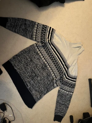 Stickad tröja med mönster i svartvitt - Stickad tröja i svart och vitt med grafiskt mönster och hög krage. Tröjan har långa ärmar, ribbade muddar och en mix av ränder och klassiska stickade detaljer. Perfekt för kalla dagar och ger en stilren look.