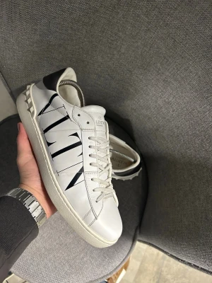 Valentino Opem - Snygga vita sneakers från Valentino Garavani med svart VLTN-logga på sidan. Skorna har vita snören, svarta detaljer vid hälen och coola nitar på baksidan. Tillverkade i slätt läder för en clean och exklusiv look.