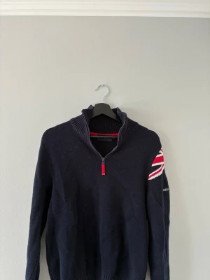 Marinblå stickad tröja Henri Lloyd - Stickad marinblå tröja från Henri Lloyd med halv dragkedja och hög krage. Union Jack-detalj i rött, vitt och blått på ena ärmen samt broderad logga. Perfekt för dig som gillar klassisk och sportig stil.