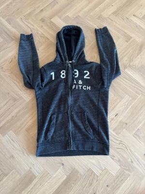 Mörkgrå hoodie från Abercrombie & Fitch - Mörkgrå ziphoodie från Abercrombie & Fitch med broderad text '1892 A&FITCH' i vitt på bröstet. Hoodien har dragkedja, två fickor fram och en stor huva. Mjuk bomullsblandning och klassisk collegekänsla.