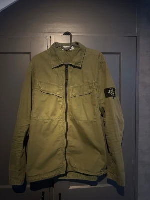 Grön overshirt från Stone Island - Overshirt från Stone Island med dragkedja framtill och två bröstfickor. Klassisk patch med logga på vänster ärm i bra skick. Tag och kasse fås med köpt på stone Island i Stockholm.
