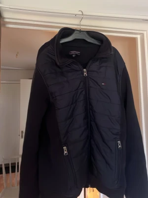 Svart hybridjacka från Tommy Hilfiger - Svart hybridjacka från Tommy Hilfiger med quiltad front, stickade ärmar och rygg. Två dragkedjefickor framtill och diskret logga på bröstet samt ärmen. Hög krage och dragkedja hela vägen. Perfekt för lager-på-lager och streetwear. Den är L men passar S.