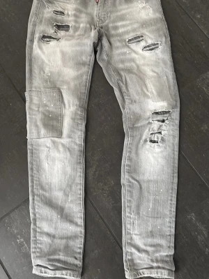 Grå slitna jeans med patchdetalj - Säljer ett par grå disquared jeans med slitningar och hål, samt en patchdetalj på vänster ben. Jeansen har raka ben och en tvättad look med vita färgstänk. 