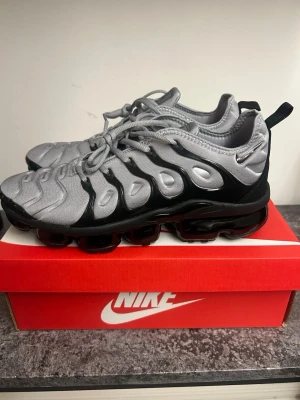 Nike Air Vapormax Plus silver/svart - Nike Air Vapormax Plus sneakers i silver och svart med futuristisk design. Ovandel i textil och syntet, tydliga vågformade detaljer och synlig Vapormax-sula i svart. Skorna har snörning och dragflik bak för enkel påtagning. Perfekta för dig som gillar streetwear.