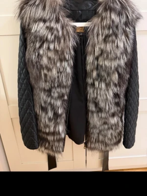 Svart pälsjacka med quiltade ärmar - Snygg svart pälsjacka med långa, quiltade ärmar och bred päls framtill i grå och svarta toner. Jackan har ett bälte i midjan för extra stil och passform. Perfekt för att hålla värmen och se trendig ut under vintern. Ärmen går att ta av så blir det en väst! Äkta päls 