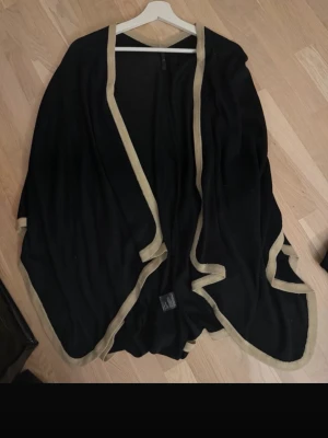 Svart poncho med beige kant - Stilren svart poncho med beige kantband runt hela plagget. Ponchon har en öppen, flödande passform och är perfekt att slänga över en outfit för extra lager. Materialet ser mjukt och följsamt ut, vilket ger en avslappnad men ändå chic vibe.
