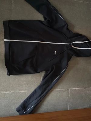 Svart hoodie med dragkedja från BOSS - Snygg svart hoodie från BOSS med hel dragkedja framtill och vit piping längs ärmarna. Liten BOSS-logga på bröstet och huva med dragsko. Tillverkad i ett mjukt och bekvämt material som passar perfekt till en avslappnad stil.