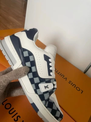 Louis Vuitton Damier sneakers blå/vit - Säljer soprena Louis Vuitton sneakers i blått och vitt med klassiskt Damier-mönster. Skorna har vita och marinblå detaljer, LV-logga på sidan och gummisula med monogramdetaljer. Materialet är en mix av läder och textil för en lyxig känsla.