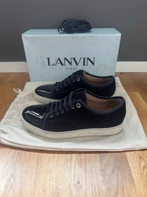 Svarta Lanvin sneakers i mocka och lack - Snygga svarta sneakers från Lanvin med ovandel i mocka och lackad tå. Skorna har vita sulor, svarta skosnören och metallfärgade öljetter. Klassisk låg modell som ger en clean och stilren look. Perfekta för dig som gillar exklusiva detaljer och premiumkänsla.