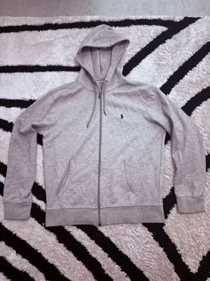Grå hoodie från Polo Ralph Lauren - En snygg grå hoodie från Polo Ralph Lauren med dragkedja och huva med snörning som inte används längre utan bara sitter i garderoben. Den är bara använt fåtals gånger. Tröjan har två fickor fram och den klassiska broderade loggan på bröstet. Perfekt för chill dagar och enkel att matcha med jeans eller joggers. Vill få bort den så fort som möjligt!!!! Kan sänkas vid snabb affär!!!! Först till kvarn