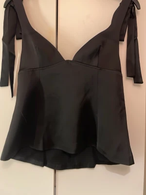 Svart satin korsett-topp med knytband - Snygg svart korsett-topp i satin med breda axelband som knyts på axlarna. Toppen har en v-ringad front och dragkedja bak. Super populär topp från Gina och är aldrig använd
