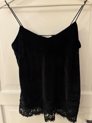 Svart linnetopp med spets från Pieces - Supersnygg svart linnetopp från Pieces med tunna axelband och dekorativ spetskant nertill. Toppen är gjord i mjuk sammet som ger en lyxig känsla och passar perfekt till olika outfits.