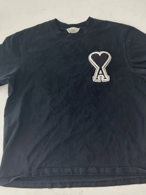Svart t-shirt från Ami Paris - Svart t-shirt från Ami Paris med en stor broderad hjärtdetalj och bokstaven A på bröstet. Klassisk rund hals och korta ärmar. Tillverkad i mjuk bomull som känns skön mot huden. Perfekt för en stilren och trendig look.