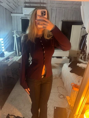 Vinröd figursydd kofta med knappar - Säljer en vinröd figursydd kofta med långa ärmar och dekorativa knappar längs hela framsidan. Koftan har en unik skärning nertill och är tillverkad i ett mjukt tyg som sitter snyggt på kroppen.