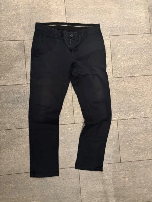 Blåa chinos med smal passform från Bläck - Snygga blåa chinos med smal passform i tailored fit modell. Byxorna har två sidofickor, en myntficka och knappgylf. Tillverkade i ett mjukt bomullsmaterial som ger en clean look. Perfekta för dig som gillar stilrena och enkla plagg.