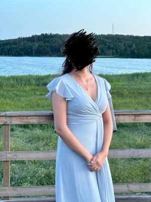 Ljusblå långklänning med volang - Säljer en elegant ljusblå långklänning med v-ringning och volangdetaljer på axlarna. Klänningen har omlott-design upptill och är ärmlös, vilket ger ett luftigt och stilrent intryck. Perfekt för sommarens tillfällen.