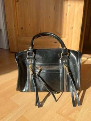 Svart handväska med gulddetaljer - Snygg svart handväska i konstläder med guldfärgade dragkedjor och dekorativa långa tofsar framtill. Väskan har två korta handtag och flera fack med dragkedja för smart förvaring. Perfekt accessoar för dig som gillar en edgy och trendig look.