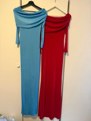 Långklänningar offshoulder blå & röd - Två eleganta långklänningar i blått och rött med offshoulder-design och trekvartsärm. Klänningarna har en åtsittande passform och är tillverkade i ett mjukt, stretchigt material som ger en snygg siluett. Perfekta för dig som vill sticka ut med färg och stil.
